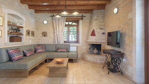 A cozy living room with a stone fireplace, a wooden ceiling, and a television mounted on the wall. - Rhadamanthus By Forest Villas (Rethymno)