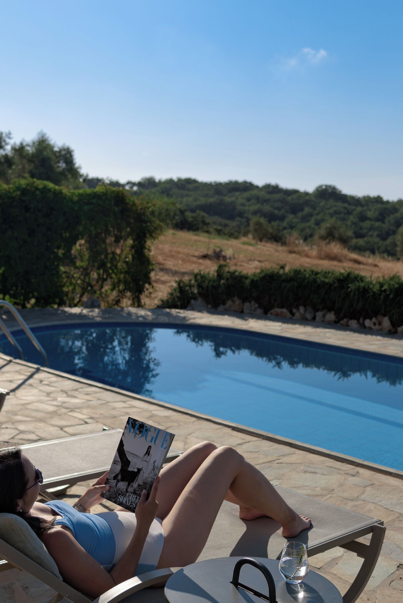 A person reclining by a poolside with a book, a glass of water, and a clear view of the pool and surrounding greenery.