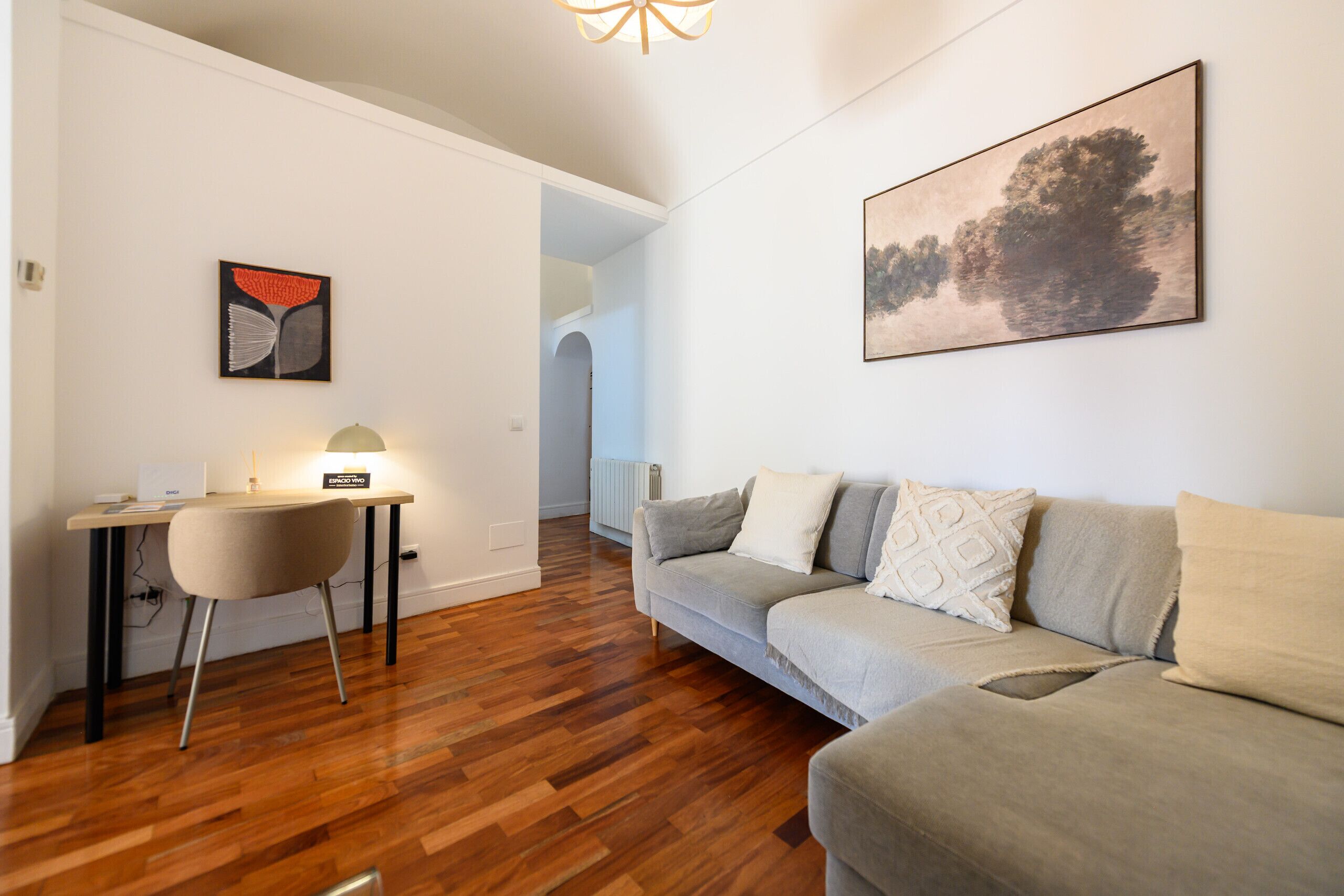 Un salón moderno con suelo de madera, un sofá gris, un escritorio con una silla y un cuadro en la pared.