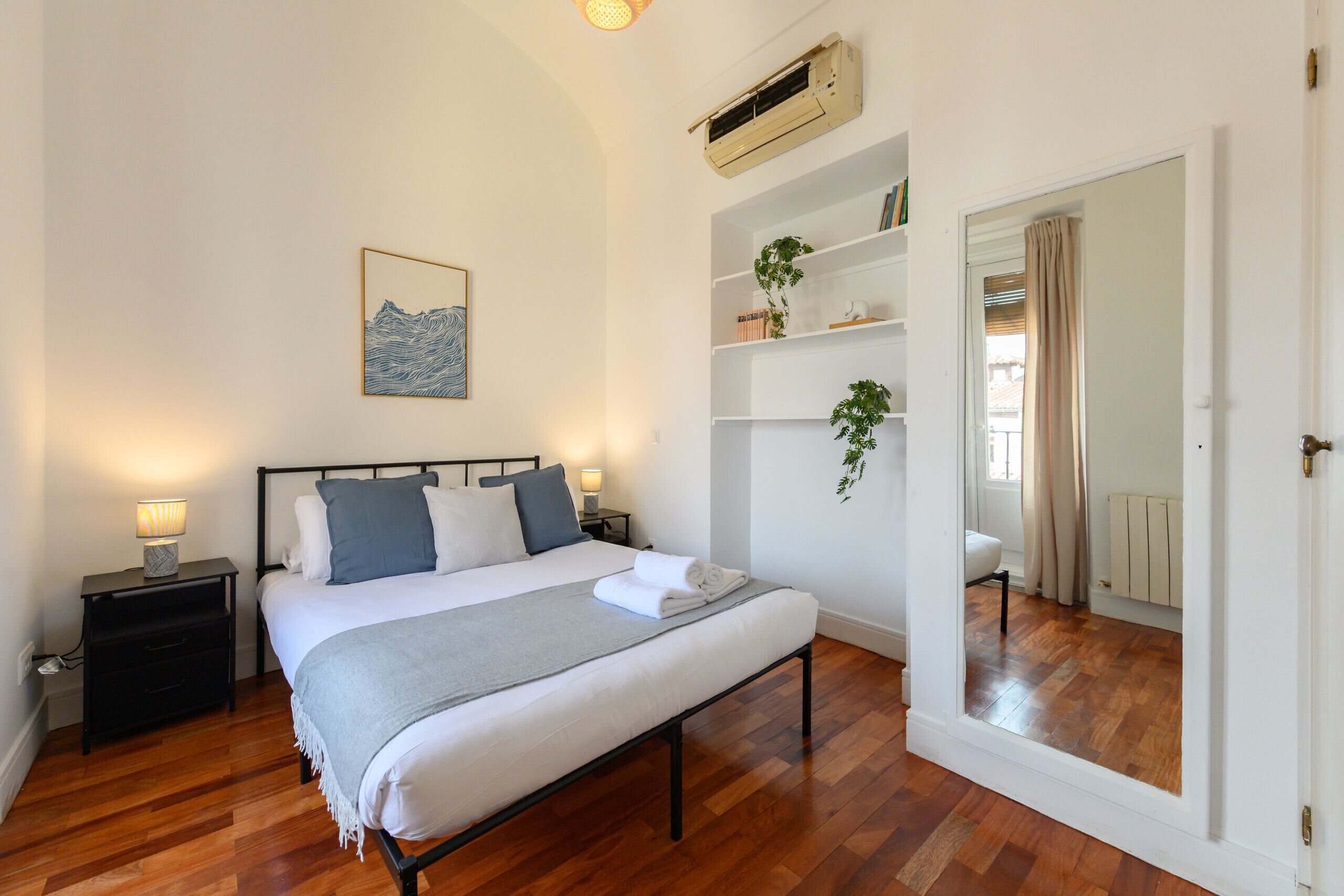 A bedroom with a bed, bedside tables, a nightstand, a wall-mounted air conditioner, a shelf with books and a plant, and a mirror.