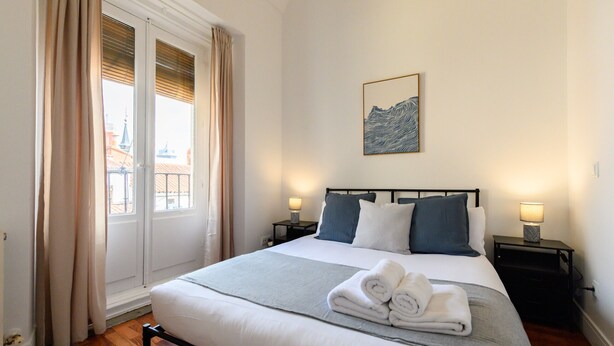 Ein ordentlich bezogenes Bett mit einer grauen Decke und zwei Kissen, ein gefaltetes Handtuch auf dem Bett und ein Bild an der Wand.