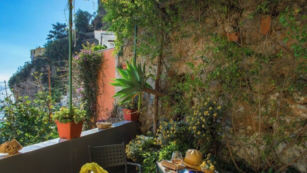 Un balcón con una silla, una mesa con un sombrero, una copa de vino y plantas en macetas.