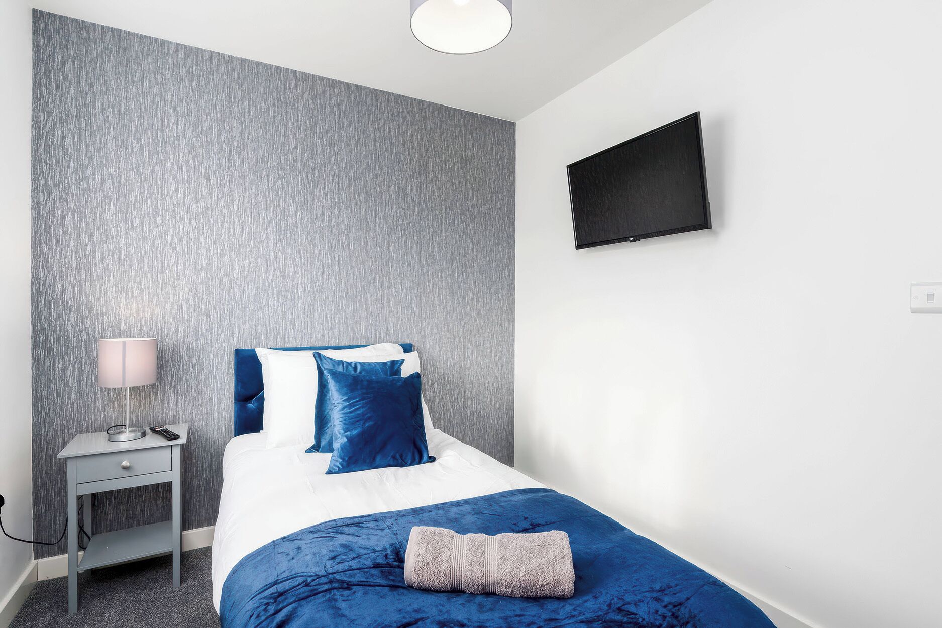 A bedroom with a bed, a bedside table, a lamp, and a wall-mounted TV.