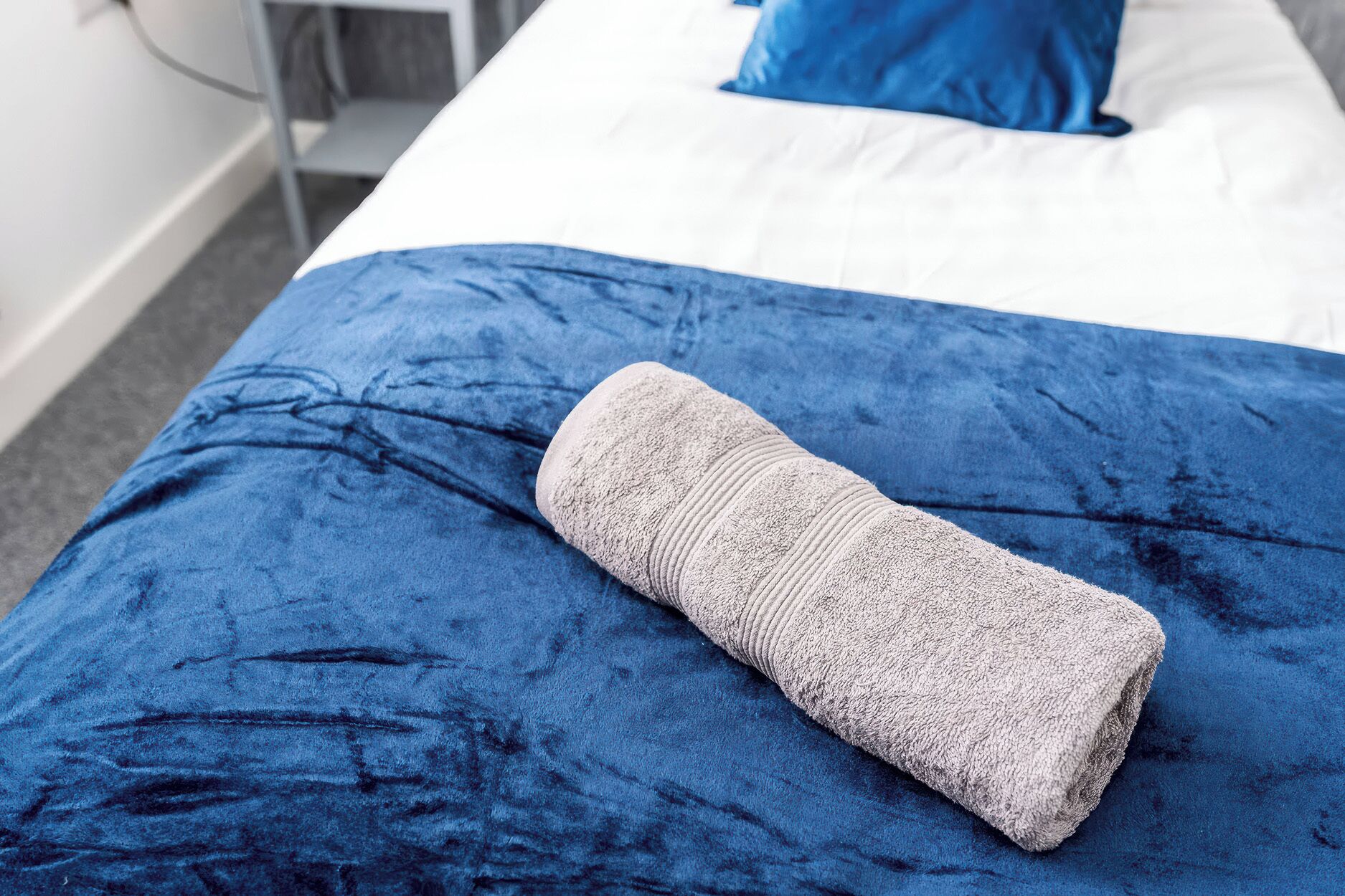 A neatly made bed with a blue bedspread and a rolled-up white towel on it.