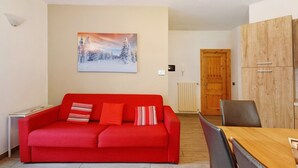 Een rode bank met gestreepte kussens, een houten eettafel en een schilderij van een besneeerd landschap aan de muur.