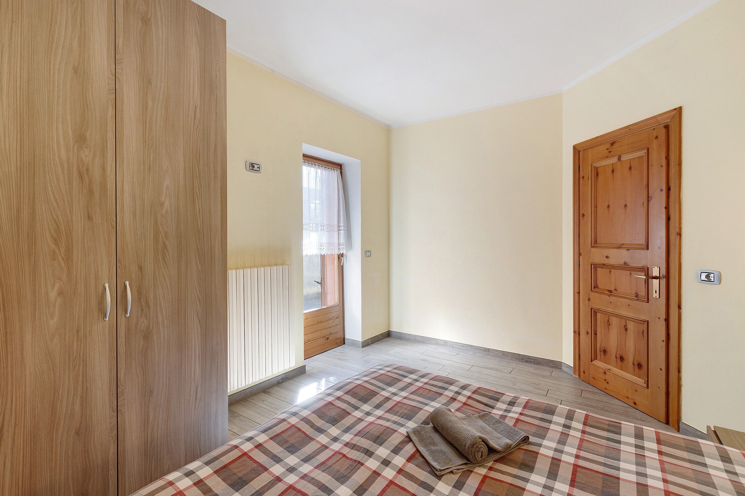 Ein Zimmer mit einem Bett, einem Holzschrank, einer Tür und einem Fenster mit Vorhang.