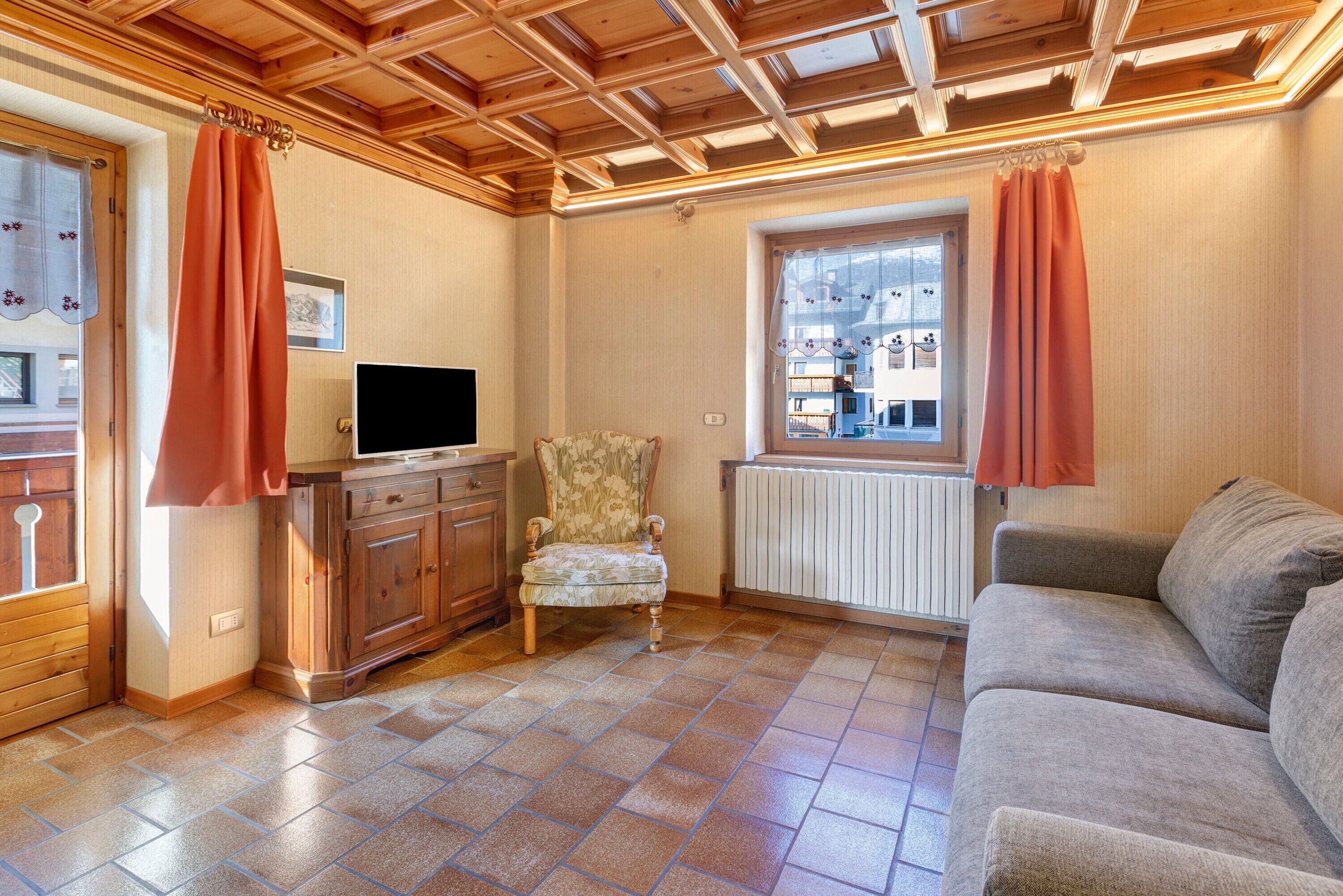 A living room with a wooden ceiling, a sofa, a chair, a TV on a cabinet, and a window with curtains.
