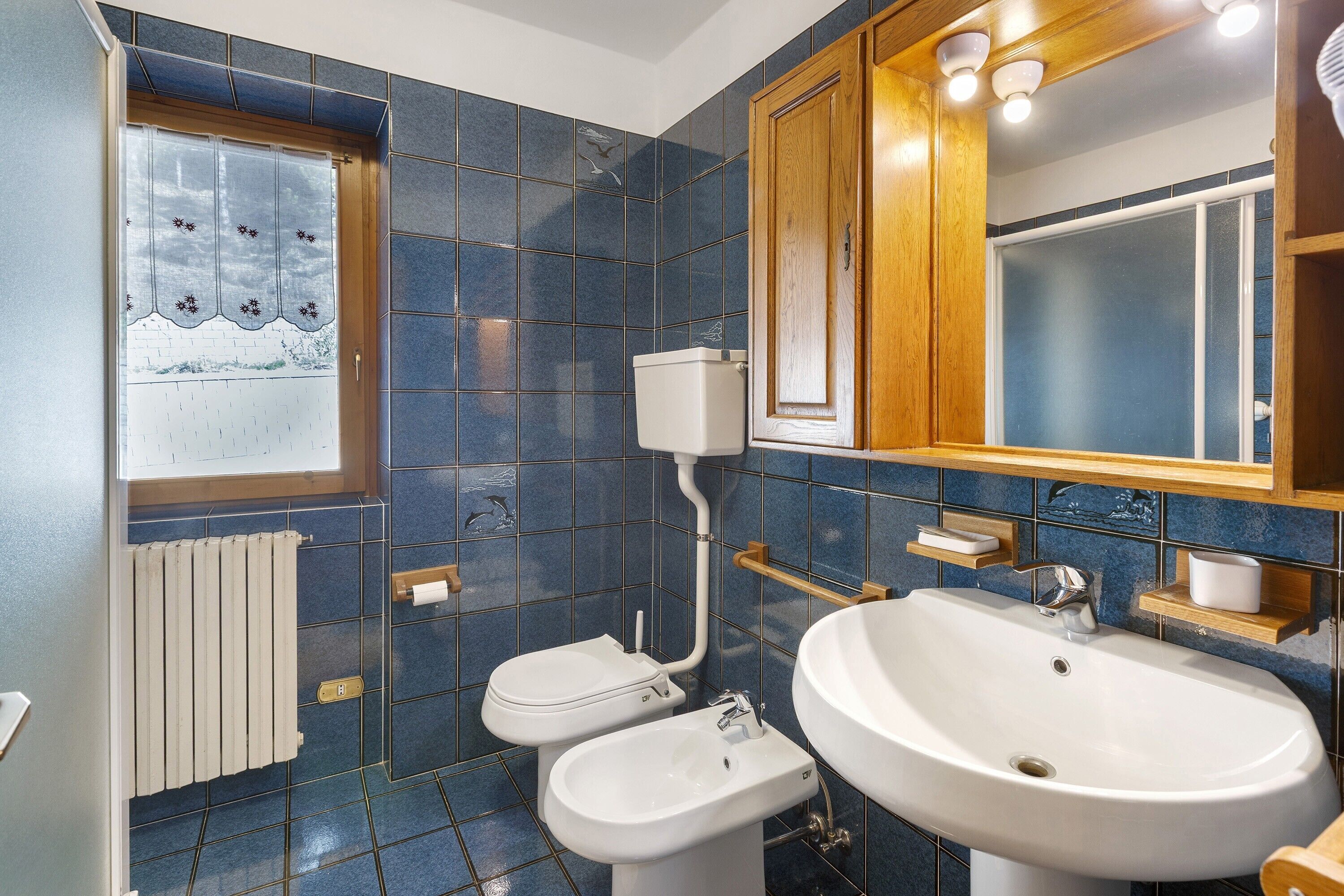 Un bagno con piastrelle blu, un water, un bidet, un lavandino, un armadietto in legno e una finestra con vista.