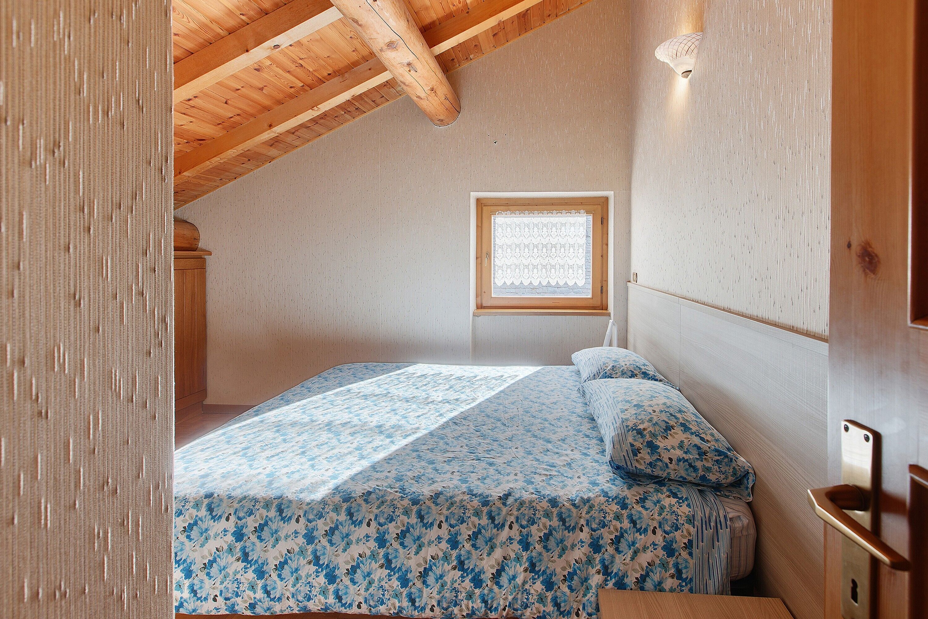 Ein Schlafzimmer mit Holzboden, einem Fenster mit blickdichter Jalousie und einem Bett mit blumigem Überwurf.