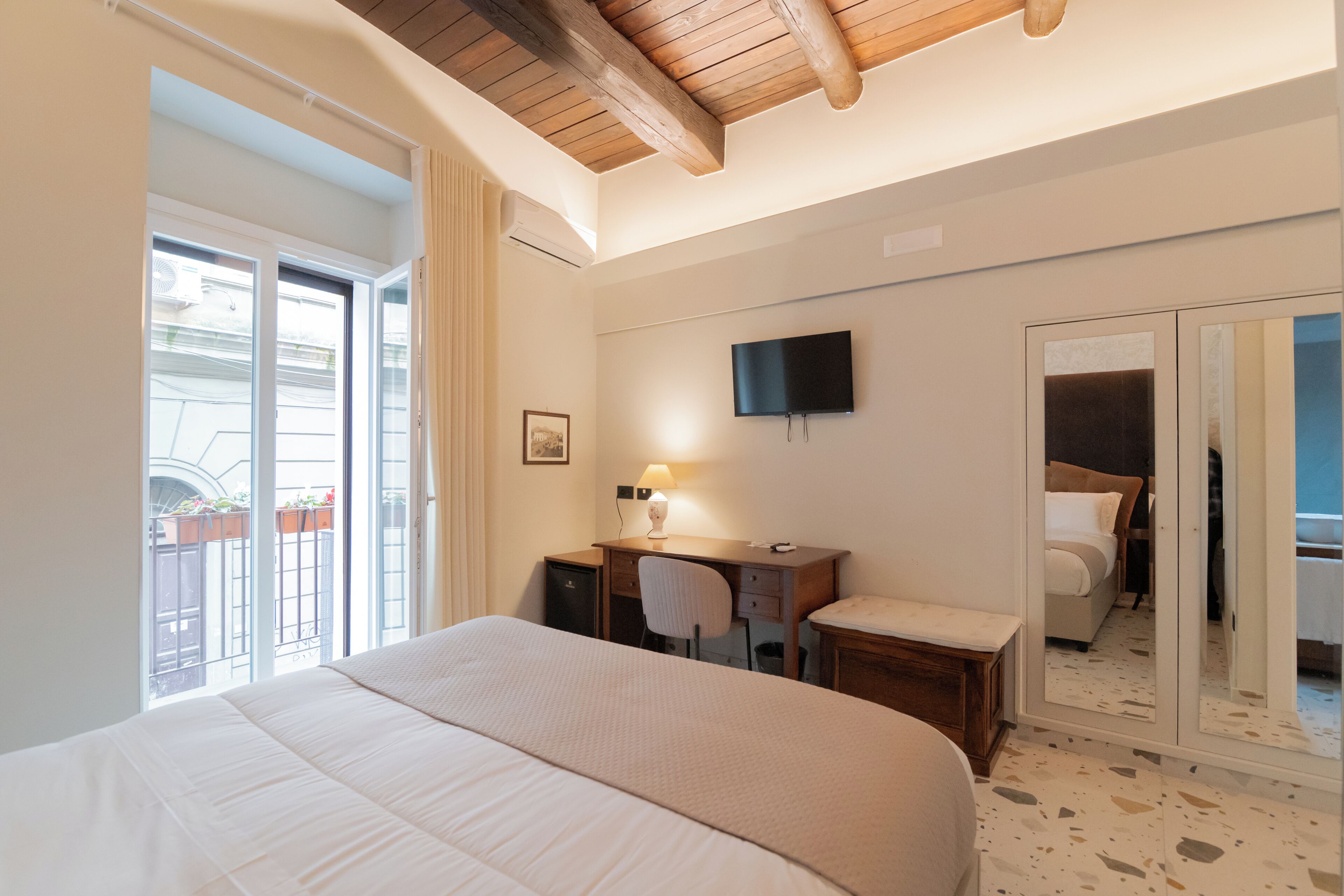 A hotel room with a large bed, a desk with a chair, a TV mounted on the wall, and a sliding door leading to a balcony.