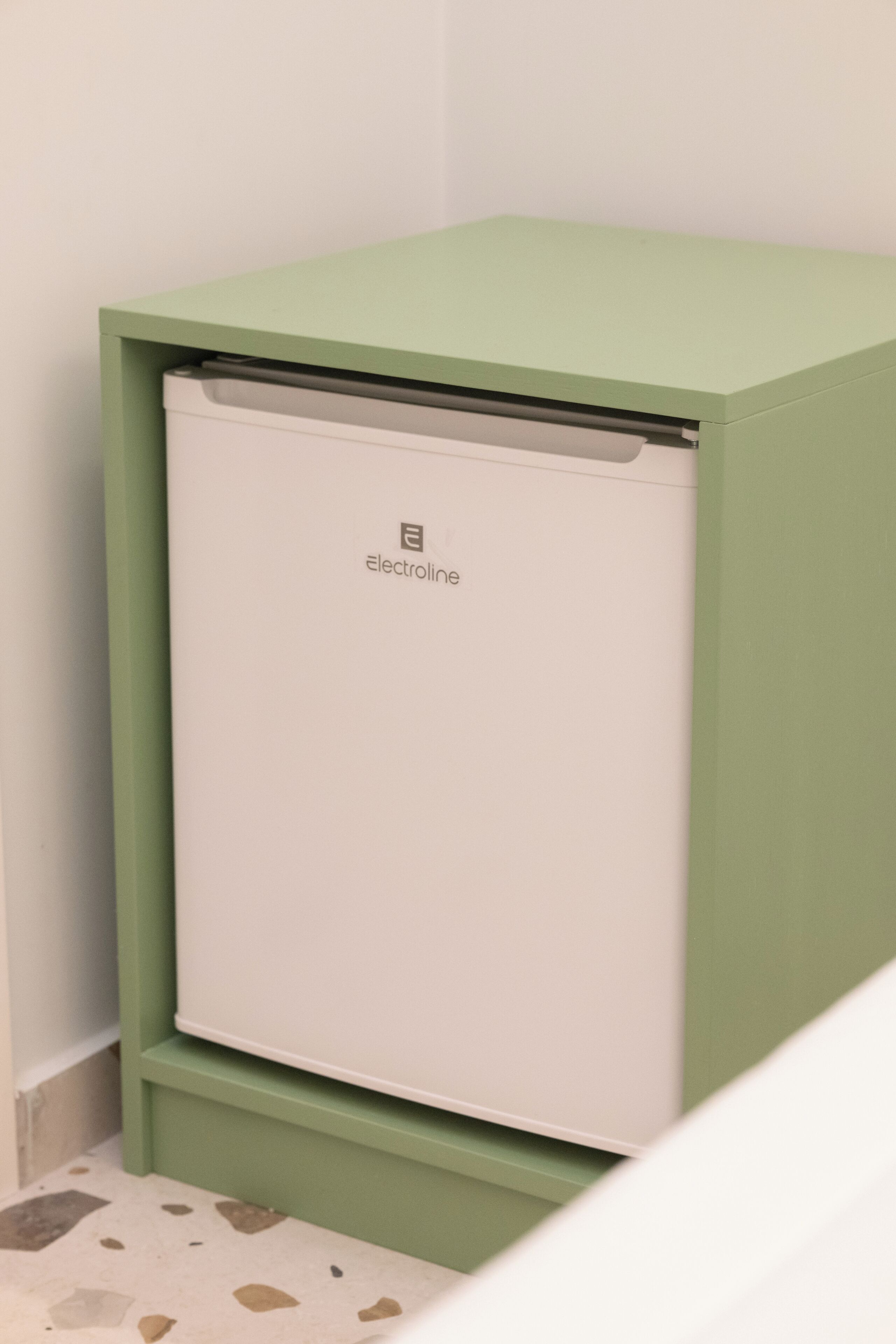 A compact, green-framed appliance, likely an air conditioner, with a white interior, placed against a wall with a speckled floor.