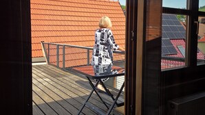 A view through a door of a wooden deck with a metal railing, a person in a patterned coat, and a red-roofed building in the background. - "Saskia" Vacation Rental with a Beautiful View of the Old Town and a Rooftop Terrace! (Hammelburg)