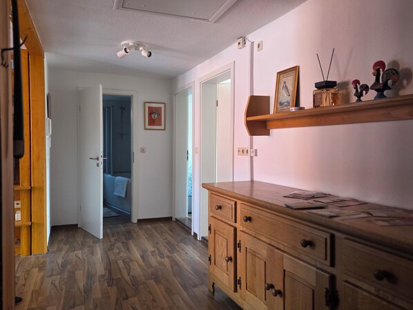A hallway with wooden flooring, a wooden cabinet with drawers, a ceiling-mounted light fixture, and a door leading to another room.
