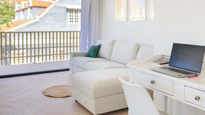 A modern living room with a white sofa, a desk with a laptop, and a view of buildings through a balcony. - Home Pietermaai in Willemstad (Willemstad)
