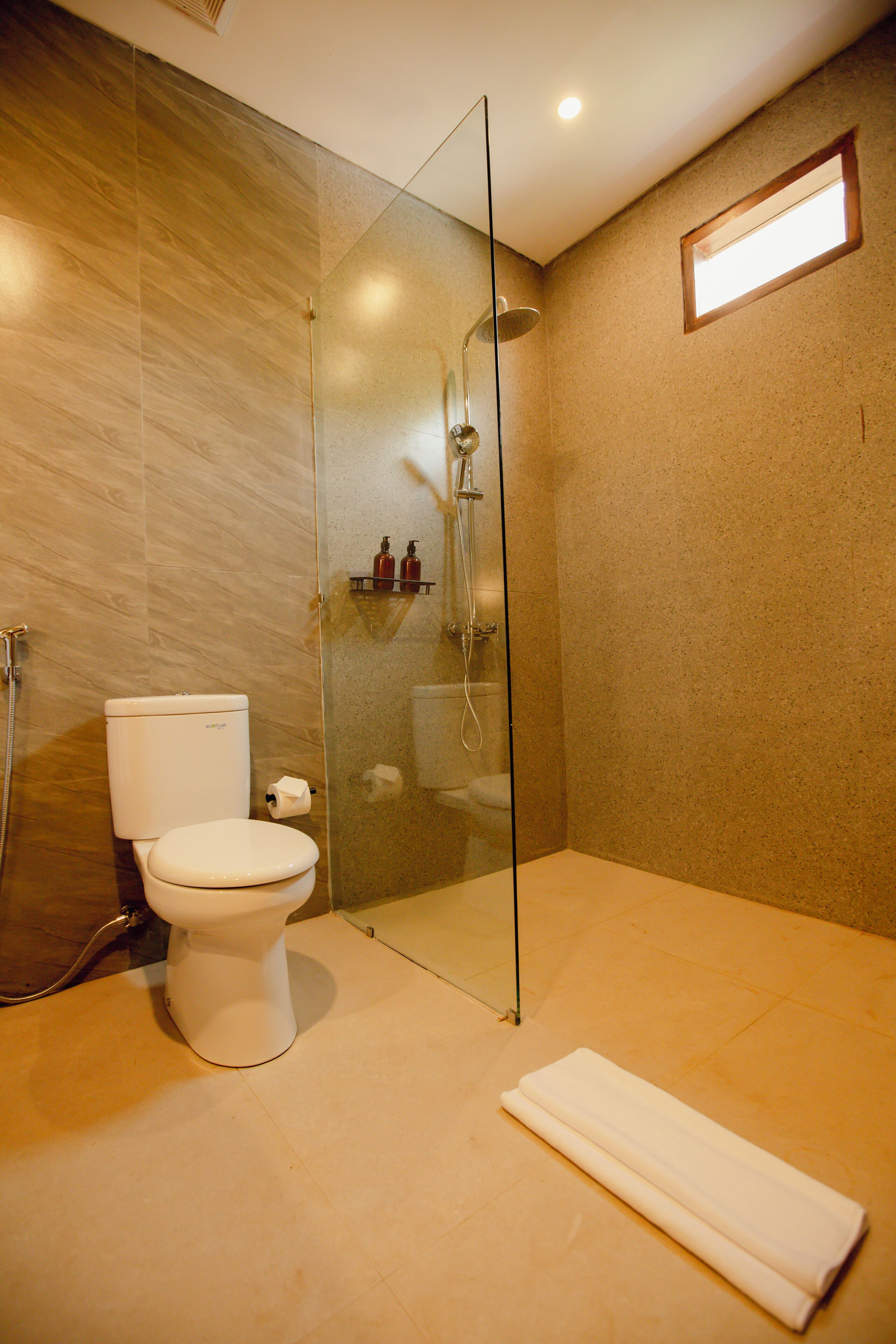 Um banheiro moderno com chuveiro separado por vidro, vaso sanitário e toalha dobrada no chão.