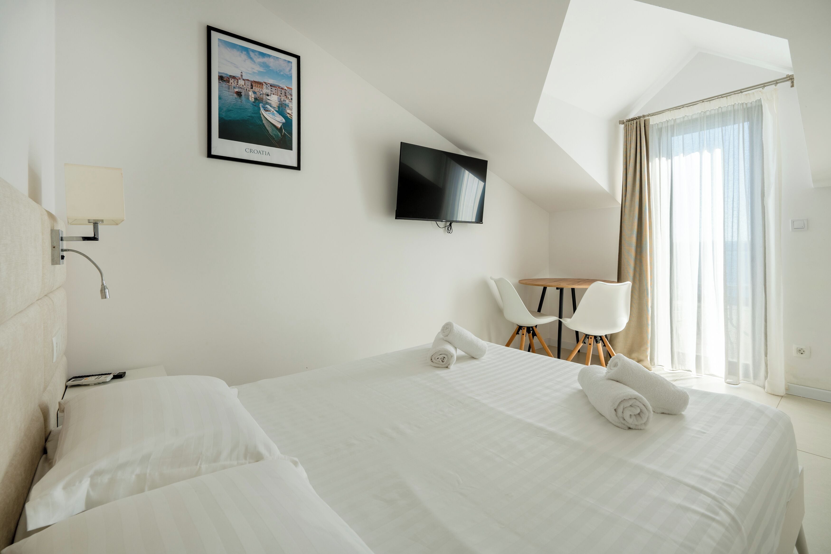 A hotel room with a large bed, a TV mounted on the wall, a desk with a chair, and a window with curtains.