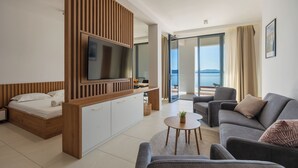 Moderni hotellihuone, jossa on sänky, televisio, sohva, pieni pöytä ja parveke, jolta on näkymä merelle.