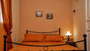 A bed with a metal frame and orange bedding, a bedside table with a lamp, and two framed pictures on the wall. - Mainades Studio - Evrypyli Serene Escape in Andros (Andros)