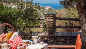 A breakfast setup with plates, cups, and a wicker basket, overlooking a coastal area with a stone wall and wooden fence. - Mainades Maisonette - Andros Seaview Escape (Andros)