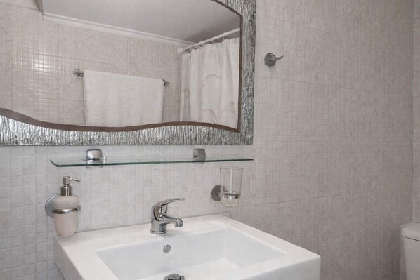 A bathroom with a white sink, a mirror with a silver frame, a glass shelf, and a shower curtain. - Absolute Beachfront Bliss in Perigiali Korinthia (Corinth)