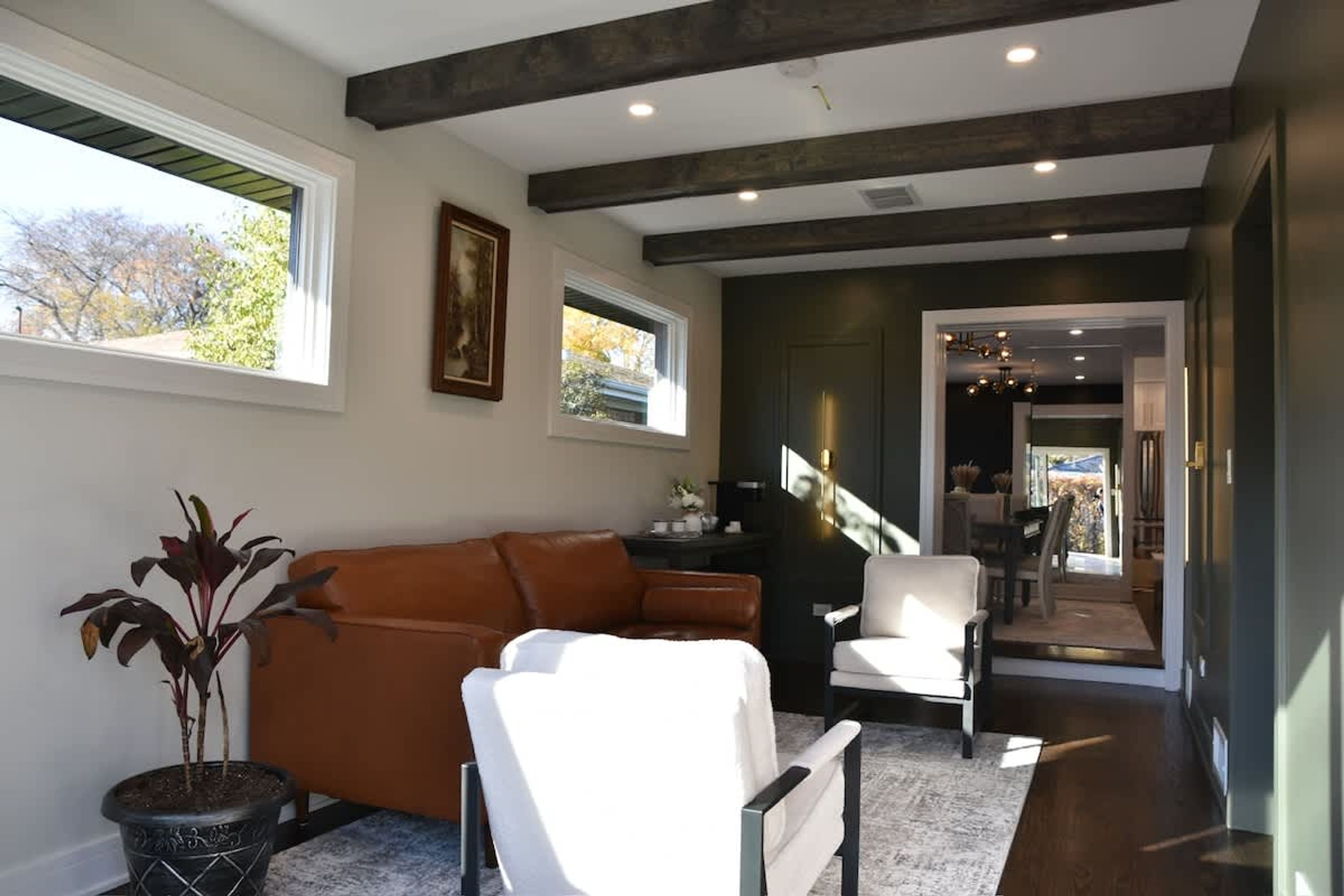 Una sala de estar moderna con un sofá de cuero marrón, un sillón blanco y una planta en maceta. Se ve una escalera que lleva a otra habitación.