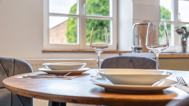 Une table à manger, dressée avec des assiettes, des couverts et des verres à vin, placée devant une fenêtre offrant une vue sur la verdure.