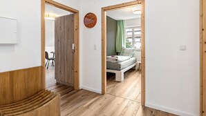 Une pièce comprenant un lit, un banc en bois, une horloge et donnant sur une autre pièce par une porte ouverte.