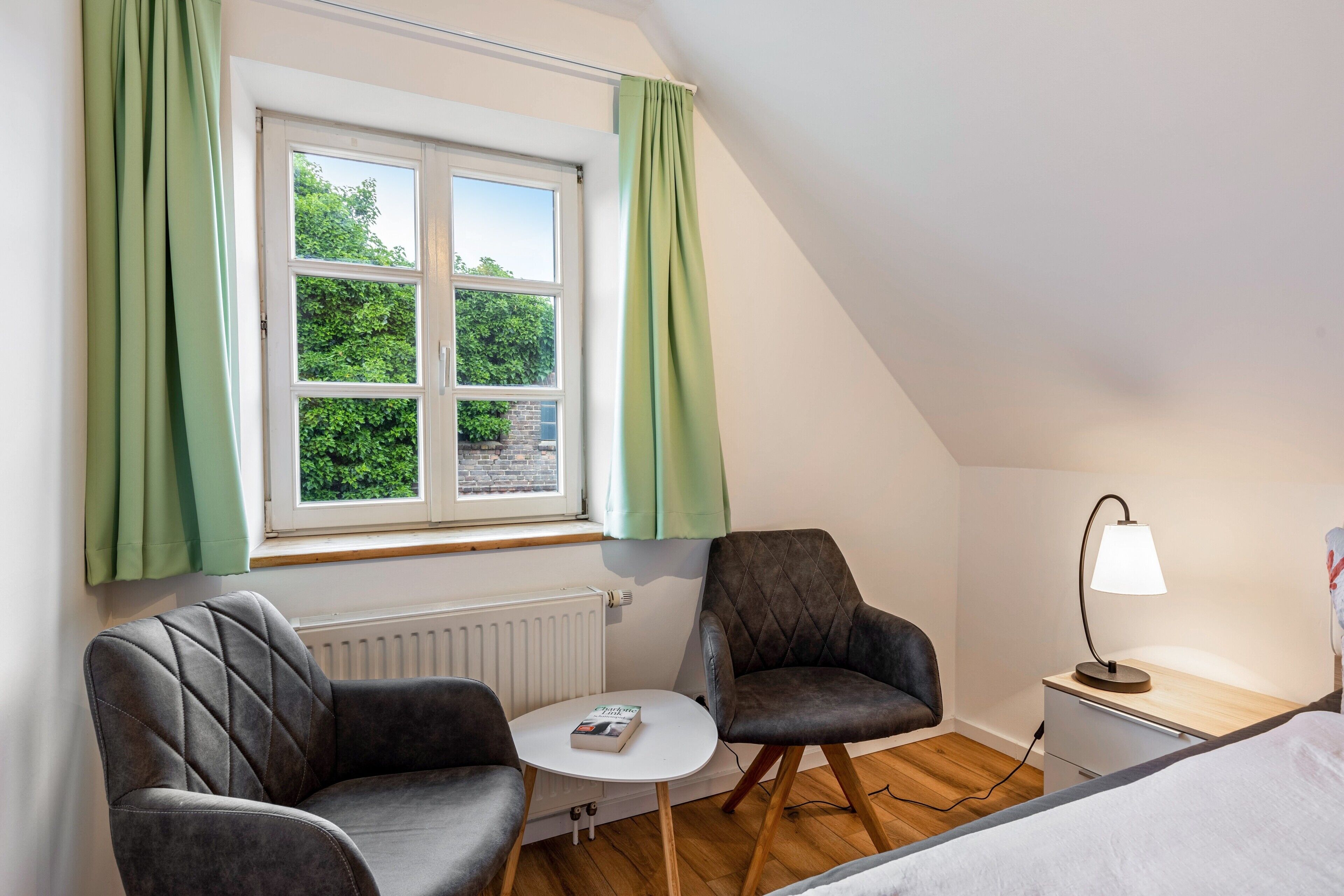 Ett rum med en säng, två fåtöljer, ett litet bord, en lampa och ett fönster med gardiner.