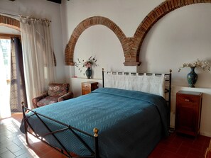 A bedroom with a bed, a chair, a nightstand, a vase with flowers, and a brick archway. - Il Peppolo Country House  (Pelago)