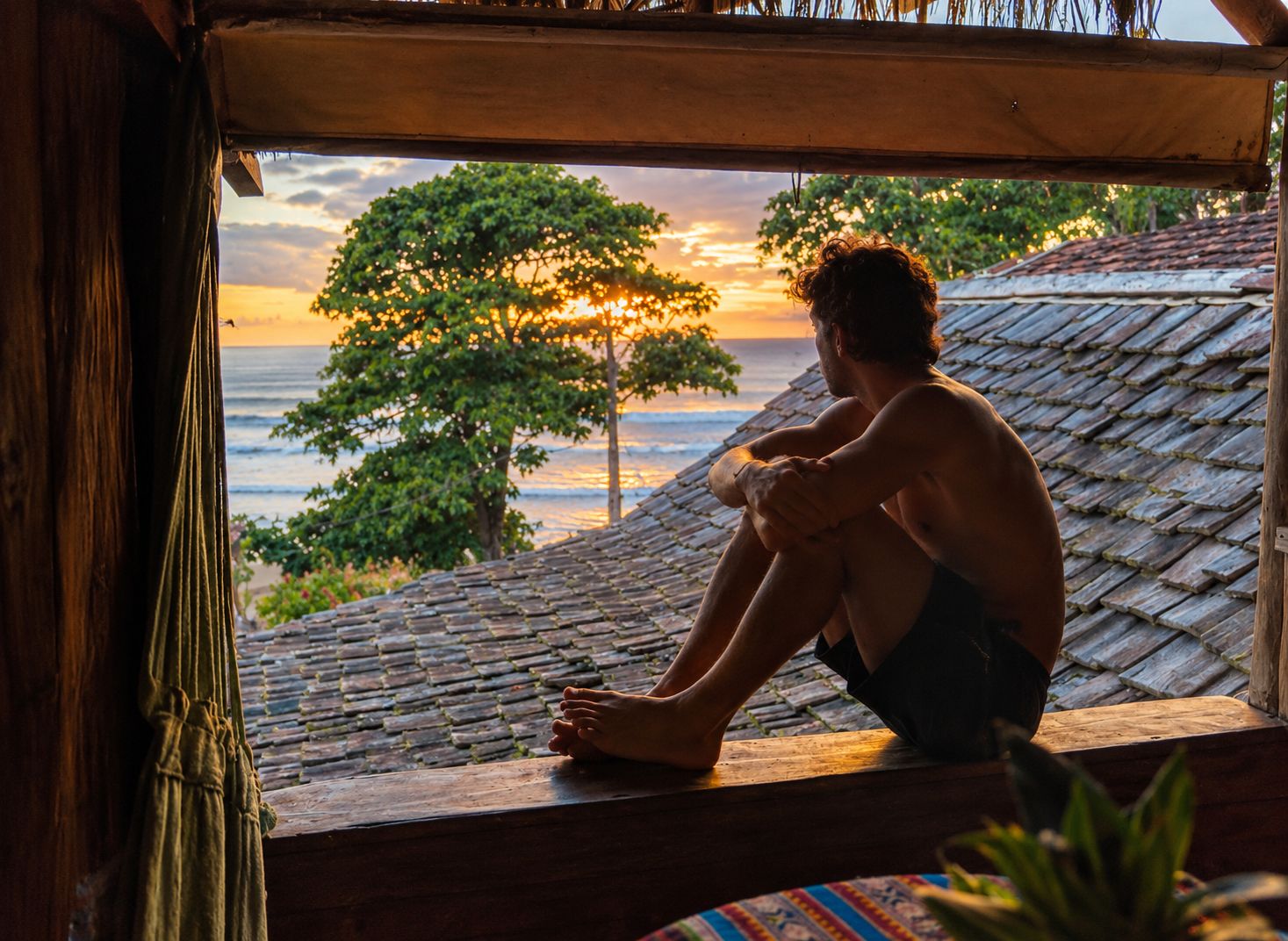 Eine Person sitzt am Fenster und blickt auf einen Sonnenuntergang, mit Blick auf das Meer und Bäume.