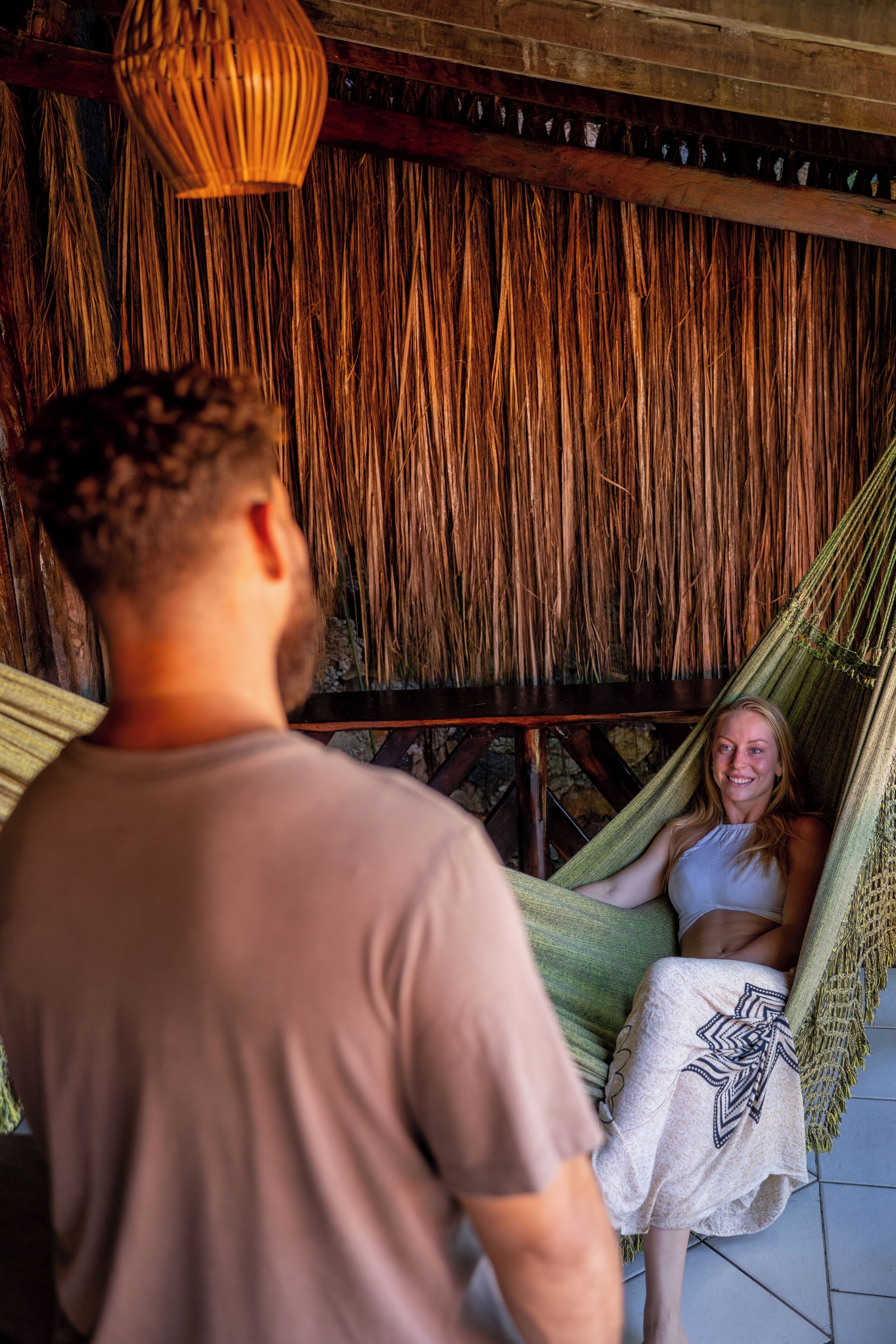 A hammock with a straw wall backdrop, a wooden table, and a hanging light fixture.