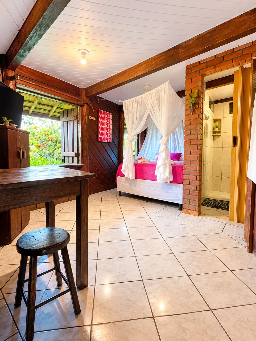 Habitación con cama bajo un dosel, una mesa de madera con un taburete y vistas al exterior con vegetación.