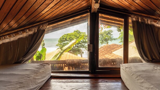 A room with a wooden ceiling, a large window, and two beds. - Beach House NATUREZA (Itacare)