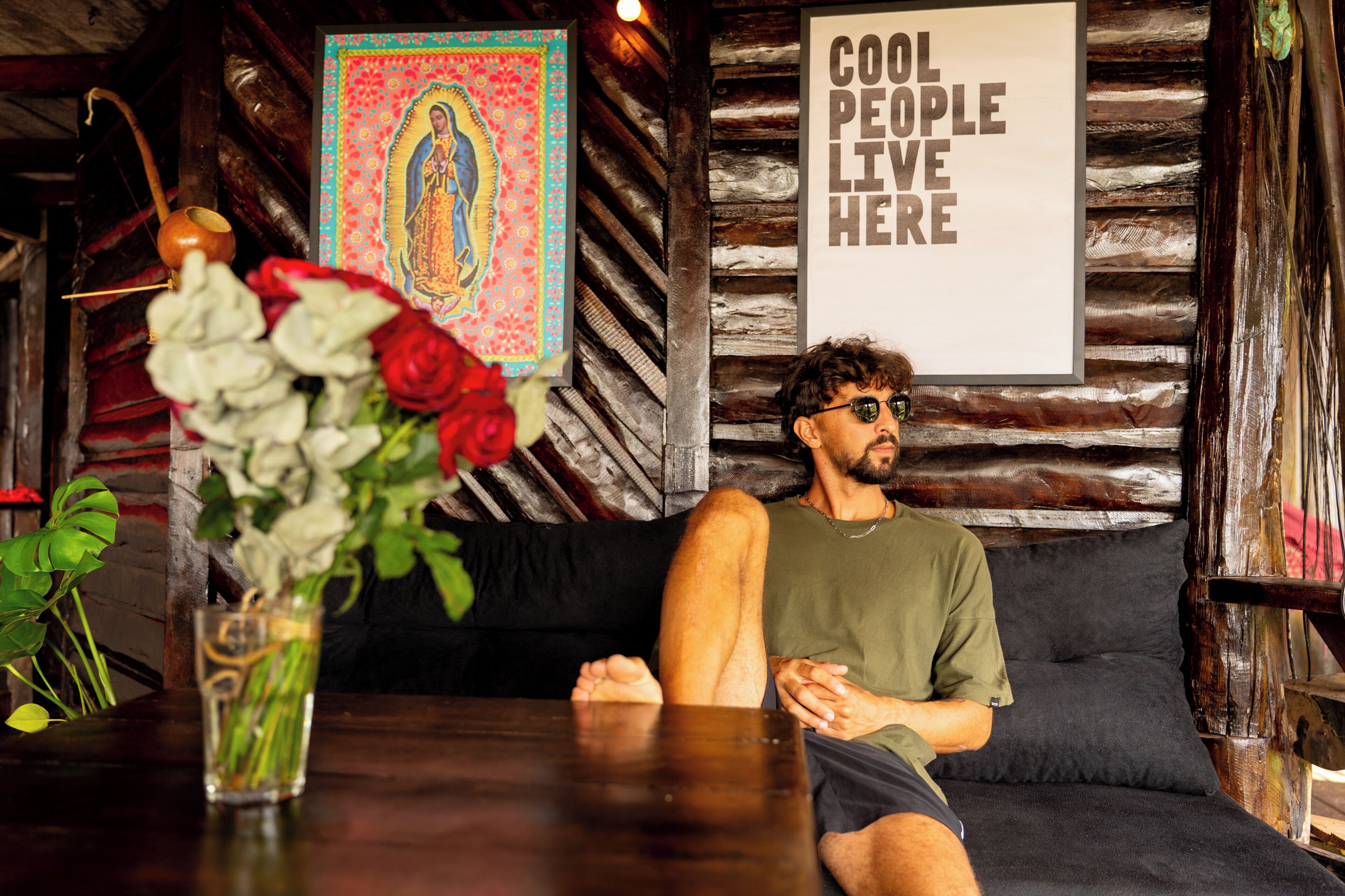 A person sitting on a couch in a rustic room with wooden walls, a poster on the wall, and a vase with flowers on a table.