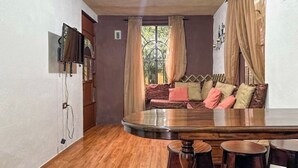 Une pièce avec un plancher en bois, un canapé avec des coussins, un téléviseur fixé au mur et une fenêtre avec des rideaux.