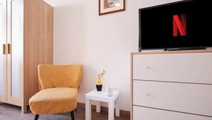 A room with a wooden wardrobe, a white dresser, a yellow armchair, a small white table with a vase, and a TV displaying the Netflix logo. - [Centocelle - Happy Domus] Oasis of the Sun (Roma)