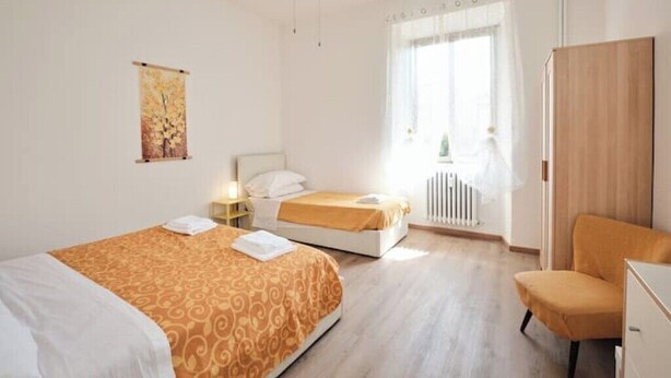A hotel room with two beds, a wooden wardrobe, a small table with a lamp, and a window with curtains. - [Centocelle - Happy Domus] Oasis of the Sun (Roma)