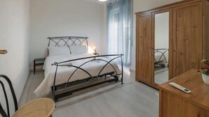 Et soveværelse med en seng med metalramme, et træskab, et lille bord med en fjernbetjening og en fletstol.
