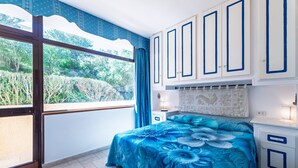 Ein Schlafzimmer mit großem Fenster, einem Bett mit Blumenmuster, weißen Schränken mit blauer Zierleiste und einem kleinen Nachttisch mit Lampe.