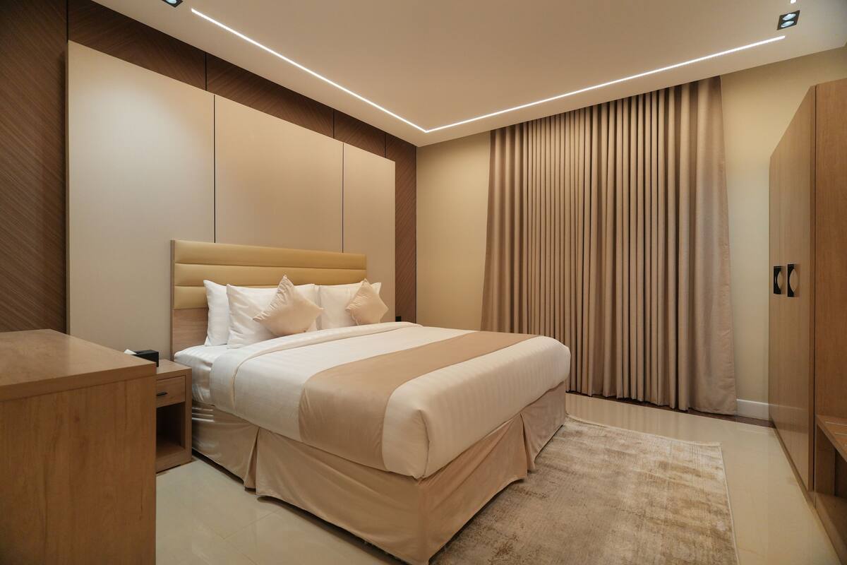 A modern hotel room with a large bed, a wooden nightstand, a desk, and a wardrobe.