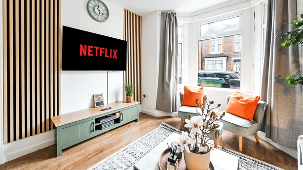 A modern living room with a flat-screen TV mounted on the wall, a wooden slat wall panel, a green entertainment unit, a window with curtains, and a view of a building outside. -  Modern Watford Hub | 4 bedroom/sleeps 8 | 25 Mins to London + 5 Mins to Center (Watford)