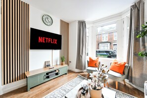 Una sala moderna con un televisor de pantalla plana montado en la pared, un panel de pared de listones de madera, un mueble de entretenimiento verde, una ventana con cortinas y vistas a un edificio.