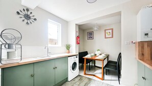 A compact kitchen with a washing machine, a dining area with a table and chairs, and a wall-mounted clock. -  Modern Watford Hub | 4 bedroom/sleeps 8 | 25 Mins to London + 5 Mins to Center (Watford)