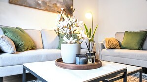 A modern living room with a sofa, a coffee table, a potted plant, and a floor lamp. -  Modern Watford Hub | 4 bedroom/sleeps 8 | 25 Mins to London + 5 Mins to Center (Watford)