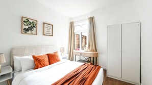 A hotel room with a bed, a nightstand, a bedside table, a wardrobe, a window with curtains, and two framed pictures on the wall. -  Modern Watford Hub | 4 bedroom/sleeps 8 | 25 Mins to London + 5 Mins to Center (Watford)