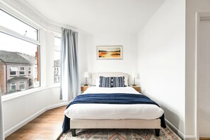 Un dormitorio con un ventanal grande, una cama con manta azul, dos mesitas de noche con lámparas y un cuadro enmarcado en la pared.