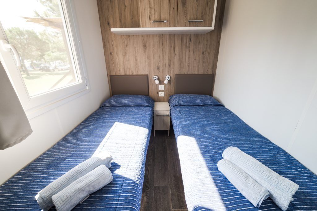 A small, compact bedroom with two single beds, a wooden headboard, a small shelf, and a window.