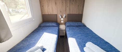 A small, compact bedroom with two single beds, a wooden headboard, a small shelf, and a window.