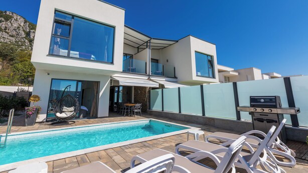 Een modern huis met twee verdiepingen, een zwembad, terrasmeubilair en een barbecueplek.