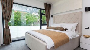 Een slaapkamer met een groot bed, een nachtkastje, een raam met gordijnen en uitzicht op groen en gebouwen.